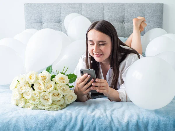 Woman in white clothes lying between white color balloons on a bed. Portrait of smiling woman using her phone with a lot of white air balloons and bouquet of roses. Happy birthday, wedding. Copy space
