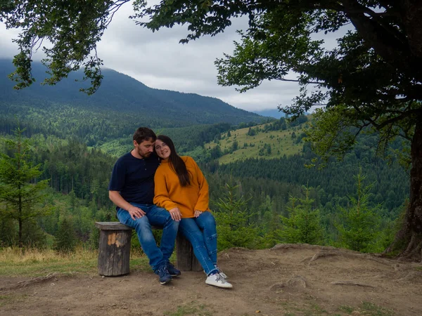 Young man and woman admiring breathtaking view while sitting on bench in the mountains. Lovely couple in casual clothes sit under large tree with forest background.