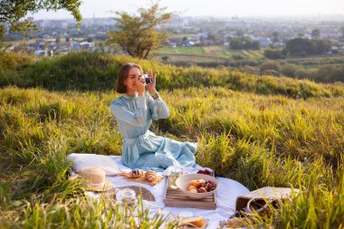 A woman in a long summer dress with short hair sitting on a white blanket with fruits and pastries and making photo of view with camera. Concept of having picnic during summer holidays or weekends. 
