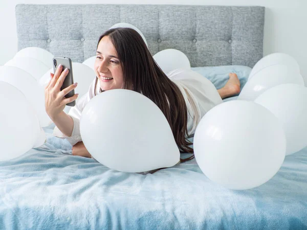 Woman in white clothes lying between white color balloons on a bed. Portrait of smiling woman using her phone with a lot of white air balloons. Happy birthday. Decoration for wedding. Copy space.