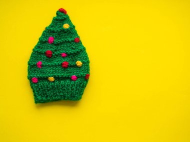 Green knitted hat in the form of a Christmas tree on a yellow background. Christmas, warming and handmade concept. New year greeting card with copy space. Knit for children.