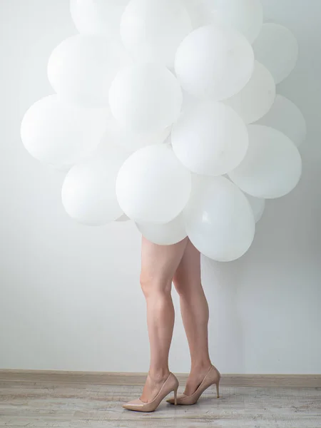 Woman in beige high heels holding white color balloons on a white wall background. Legs and a lot of white air balloons. Happy birthday anniversary. White decoration. Copy space