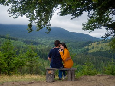 Young man and woman admiring breathtaking view while sitting on bench in the mountains. Lovely couple in casual clothes sit under large tree with forest background.