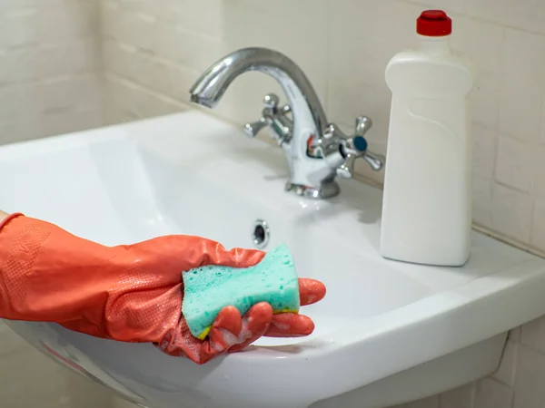 Cleaning bathroom sink and faucet with detergent in orange rubber gloves with green sponge. Housework, cleaning, hygiene home concept. Person hand holding a mint sponge with white foam.