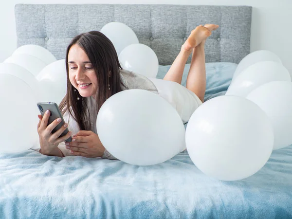 Woman in white clothes lying between white color balloons on a bed. Portrait of smiling woman using her phone with a lot of white air balloons. Happy birthday. Decoration for wedding. Copy space.