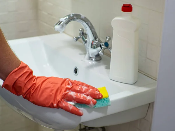 Cleaning bathroom sink and faucet with detergent in orange rubber gloves with green sponge. Housework, cleaning, hygiene home concept. Hand holds a mint sponge with white foam and scrubs the sink.