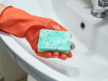Cleaning bathroom sink and faucet with detergent in orange rubber gloves with green sponge. Housework, cleaning, hygiene home concept. Person hand holding a mint sponge with white foam.