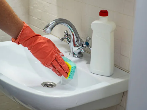 Cleaning bathroom sink and faucet with detergent in orange rubber gloves with green sponge. Housework, cleaning, hygiene home concept. Hand holds a mint sponge with white foam and scrubs the sink.