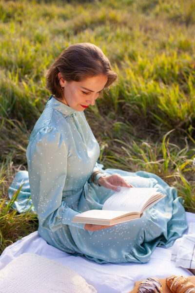 A woman in a long summer dress with short hair sitting on a white blanket with fruits and pastries and reading the book. Concept of having picnic in a city park during summer holidays or weekends. 
