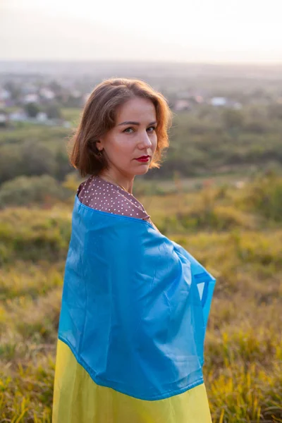 Sad Woman covering herself a yellow and blue flag of Ukraine on a background of sunset. Ukrainians against the war. Independence Day. Victim of an invasion. Rear view.