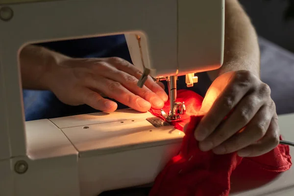 Hands of experienced worker in the handmade industry, sewing red fabric on a sewing machine, the production of Santa red hat. Homemade sewing. Working tailor. Christmas hat. Close up.