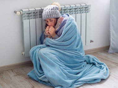 Evde akünün yanında oturan ve ısınmaya çalışan bir kadın. Sıcak giyimli kadın ısıtmanın yüksek fiyatına üzülüyor. Pahalı enerji kaynakları, Avrupa 'da enerji krizi.
