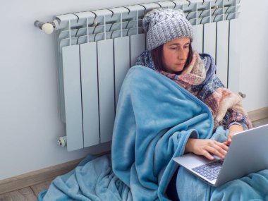 Kadın evde akünün yanında oturuyor ve dizüstü bilgisayarla çalışıyor. Sıcak giyimli kadın ısıtmanın yüksek fiyatına üzülüyor. Pahalı enerji kaynakları, Avrupa 'da enerji krizi. Çevrimiçi eğitim, iş