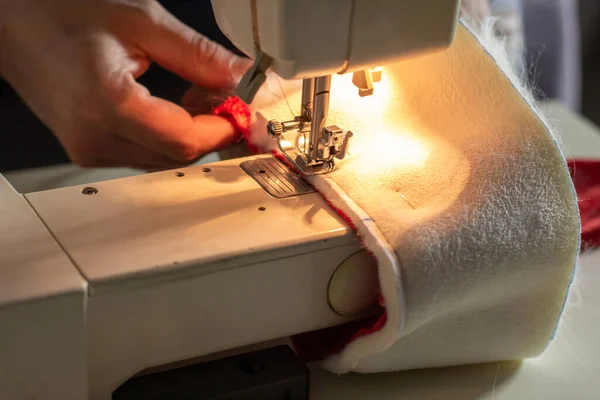 Hands of experienced worker in the handmade industry, sewing red and white fabric on a sewing machine, the production of Santa red hat. Homemade sewing. Working tailor. Christmas hat. Close up.