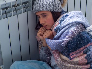 Evde akünün yanında oturan ve ısınmaya çalışan bir kadın. Sıcak giyimli kadın ısıtmanın yüksek fiyatına üzülüyor. Pahalı enerji kaynakları, Avrupa 'da enerji krizi.
