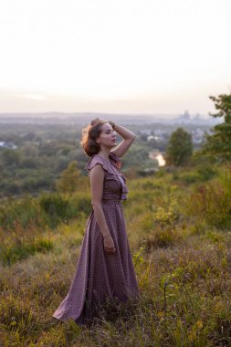 Uzun kahverengi elbiseli, gülümseyen güzel bir kız çimlerin üzerinde duruyor. Mutlu kadın gün batımında nehre bakan bir tepede yürüyor. Yaz tatillerinde ya da hafta sonlarında parkta dinlenme kavramı