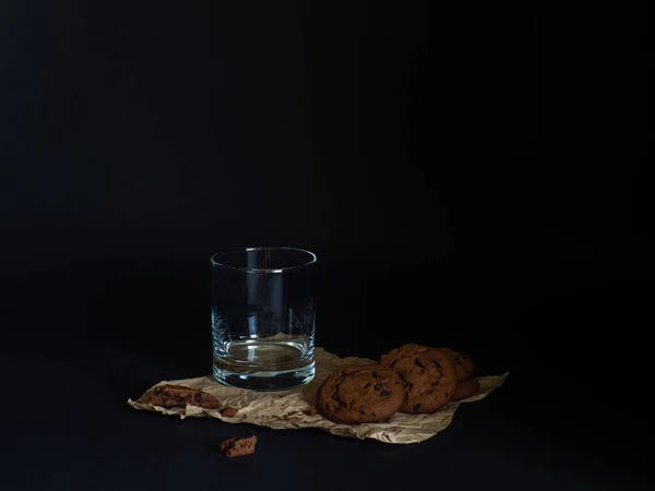 stock image Cookies made from cocoa and with pieces of chocolate inside and empty glass on a baking paper on a black background. Copy space