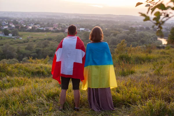 ウクライナとスイスの国旗に覆われたカップル ウクライナとスイス連邦の友好の概念 二国間の愛 男と女は日没を背景に異なる旗をカバー — ストック写真