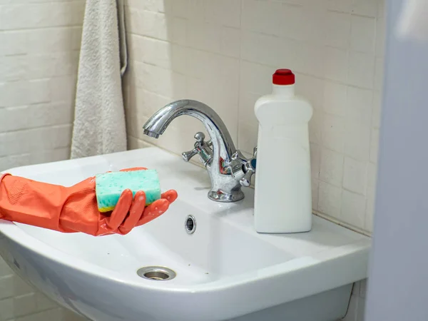 Cleaning bathroom sink and faucet with detergent in orange rubber gloves with green sponge. Housework, cleaning, hygiene home concept. Person hand holding a mint sponge in front of bathroom sink.