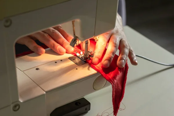 Hands of experienced worker in the handmade industry, sewing red fabric on a sewing machine, the production of Santa red hat. Homemade sewing. Working tailor. Christmas hat. Close up.