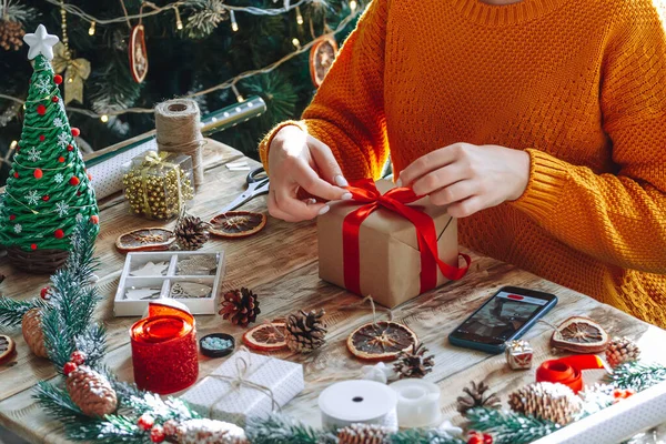 Cep telefonundaki online talimatları izleyen genç bir kadın noel hediyesi paketliyor.