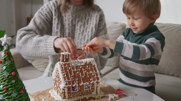 Annesiyle birlikte Noel 'de zencefilli kurabiye evi dekore eden küçük çocuk, Noel ve Noel arifesinde aile aktiviteleri ve gelenekler — Stok video