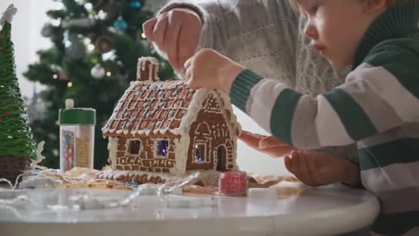 Niño pequeño con madre decorando casa de jengibre de Navidad juntos, actividades familiares y tradiciones en Navidad y Nochevieja — Vídeos de Stock