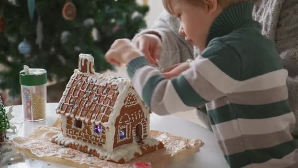 Niño pequeño con madre decorando casa de jengibre de Navidad juntos, actividades familiares y tradiciones en Navidad y Nochevieja — Vídeo de stock
