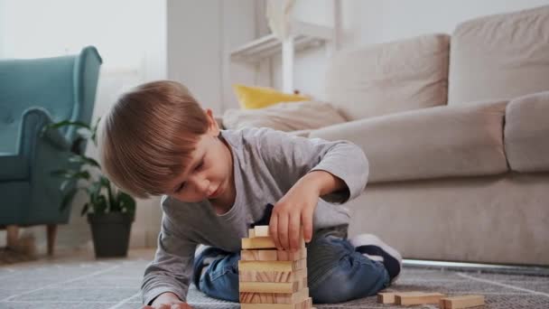 Παιδί χτίζει ένα ξύλινο πύργο για να παίζει με ξύλινα μπλοκ κάθεται στο πάτωμα στο σπίτι — Αρχείο Βίντεο