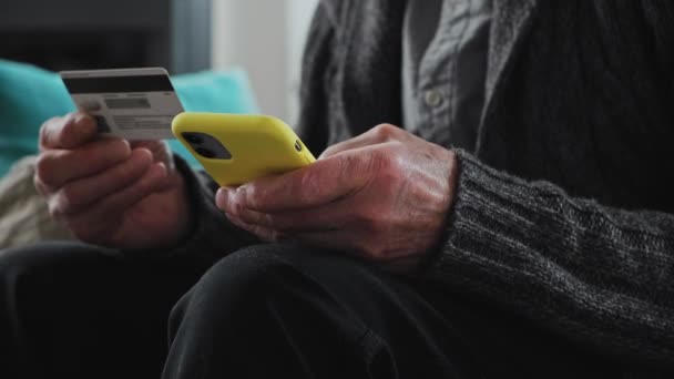 Senior man voert credit card gegevens in mobiele telefoon voor online winkelen, zittend op de bank thuis, close-up — Stockvideo