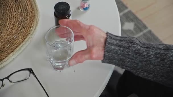 Ηλικιωμένος ανώτερος πίνει ένα ποτήρι νερό, ένα μπουκάλι χάπια είναι στο τραπέζι, στο σπίτι — Αρχείο Βίντεο