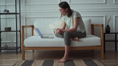Genç bir kadın evinde kanepede otururken dizüstü bilgisayar kullanıyor, rahatlıyor ve video seyrediyor.