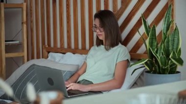 Dizüstü bilgisayar kullanan, daktilo kullanan, yatağa uzanan, uzaktan kumandalı, evde çalışan genç bir kadın.