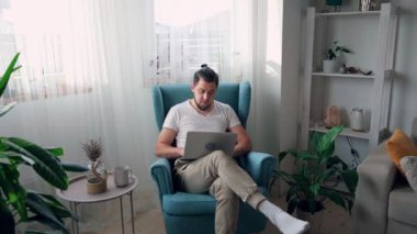 Genç adam dizüstü bilgisayar kullanıyor. Yazıyor ve çalışıyor. Evdeki koltukta oturuyor.