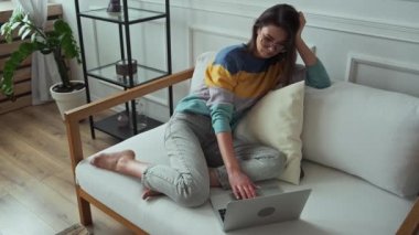 Genç bir kadın dizüstü bilgisayar kullanıyor, daktilo kullanıyor, dokunmatik ped kullanıyor, evinde oturuyor, koltukta oturuyor, uzaktan çalışıyor, evde çalışıyor.