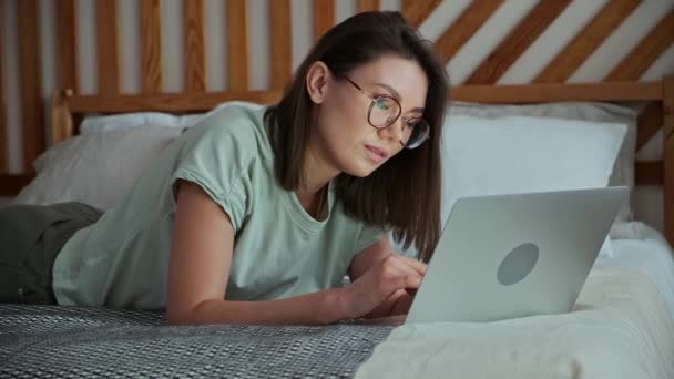 Mujer joven usando el ordenador portátil, mecanografía, acostado en la cama, trabajo remoto, trabajo en casa — Vídeos de Stock