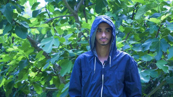 年轻的阿拉伯男子 身穿蓝色防水外套 头戴头罩 在绿林中向侧面张望 严重的男性本性 — 图库照片