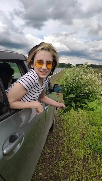Una chica en gafas amarillas, con un ramo de flores, mira hacia fuera del coche. Vacaciones. Viajes. Fotos de stock libres de derechos