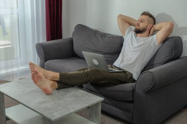 Genç gülümseyen adam evde dizüstü bilgisayar ve video konferans ile çalışırken rahatlıyor. Genç yaratıcı adam akıllı telefon kullanarak ekran mesajına bakıyor. Evden çalışıyor.