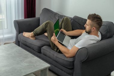 Genç gülümseyen adam evde dizüstü bilgisayar ve video konferans ile çalışırken rahatlıyor. Genç yaratıcı adam akıllı telefon kullanarak ekran mesajına bakıyor. Evden çalışıyor.