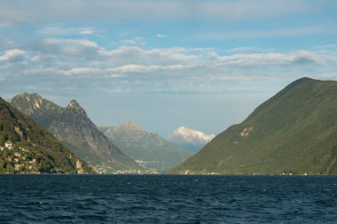 Lugano, İsviçre - 6 Ekim 2021: Lago di Lugano çevresindeki tepeler ve dağlarla öğleden sonra güneş ışığı altında.