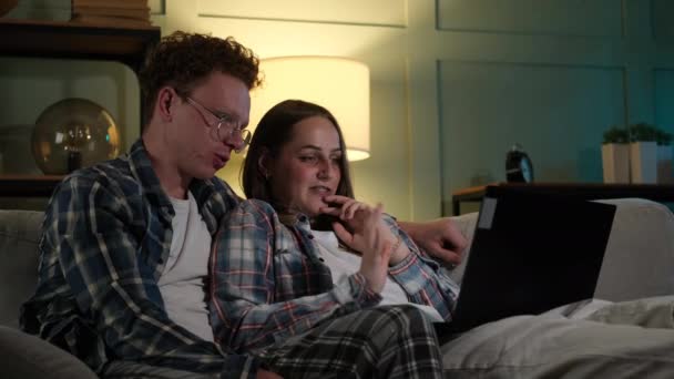 Пара сидящих на диване и наблюдающих за чем-то на ноутбуке — стоковое видео