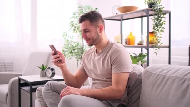Щасливий чоловік сидить на дивані за допомогою смартфона — стокове відео