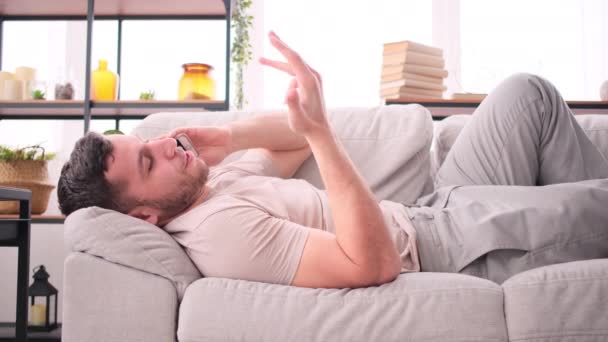 Мужчина разговаривает по мобильному телефону, лежит дома на диване — стоковое видео