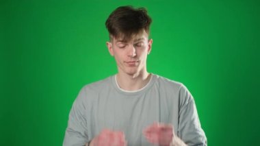 Gri tişörtlü, kafası karışmış bir adam elinle gözlerini kapatıyor ve görülmekten tiksindiğini söylüyor. Yeşil arka planda izole edilmiş kapalı stüdyo çekimi