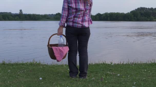 Жінка стоїть на березі озера з вінтажним кошиком для пікніка — стокове відео