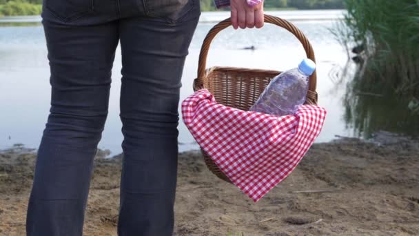 Woman stands by lake shore with vintage picnic basket — 图库视频影像