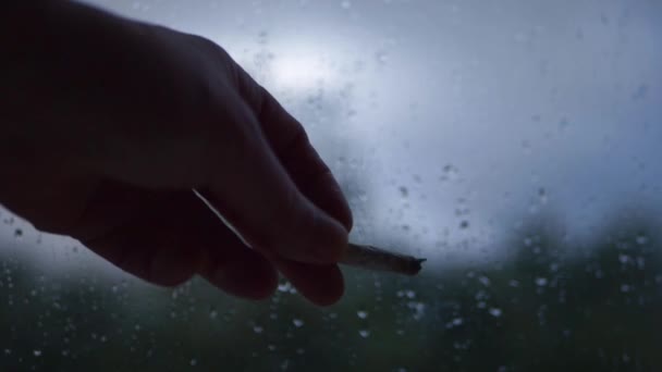 Hände des Rauchers vor dem Fenster, als es draußen an einem dunklen Regentag regnet — Stockvideo