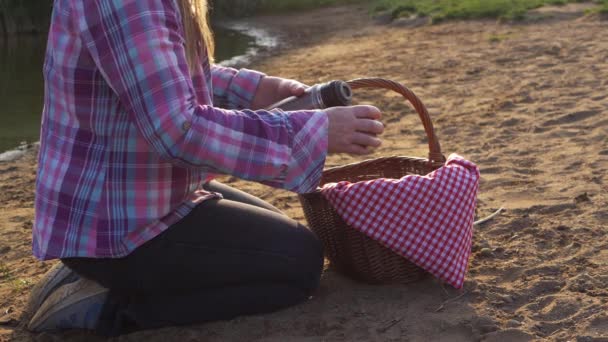 Жінка з кошиком для пікніка п'є каву з термос колби біля озера — стокове відео