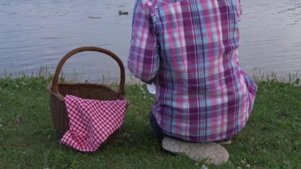 Женщина с корзиной для пикника сидит у озера — стоковое видео